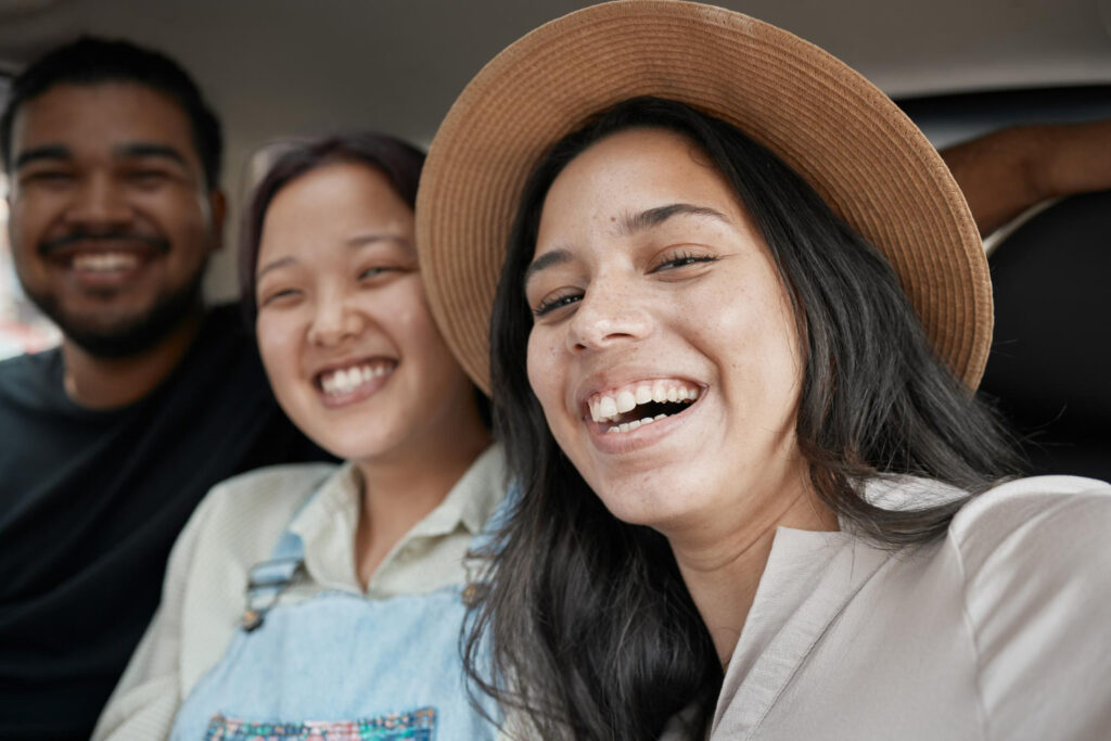 The image captures three friends sharing a joyful moment together, likely inside a vehicle, judging from the seat belts and car interior visible. The group consists of a man and two women, one of whom is wearing a stylish hat, all beaming with wide smiles. Their expressions and casual attire convey a sense of comfort and happiness in each other's company, highlighting a candid and lighthearted interaction. Such images are great for illustrating themes of friendship, diversity, and leisure activities in various media projects.